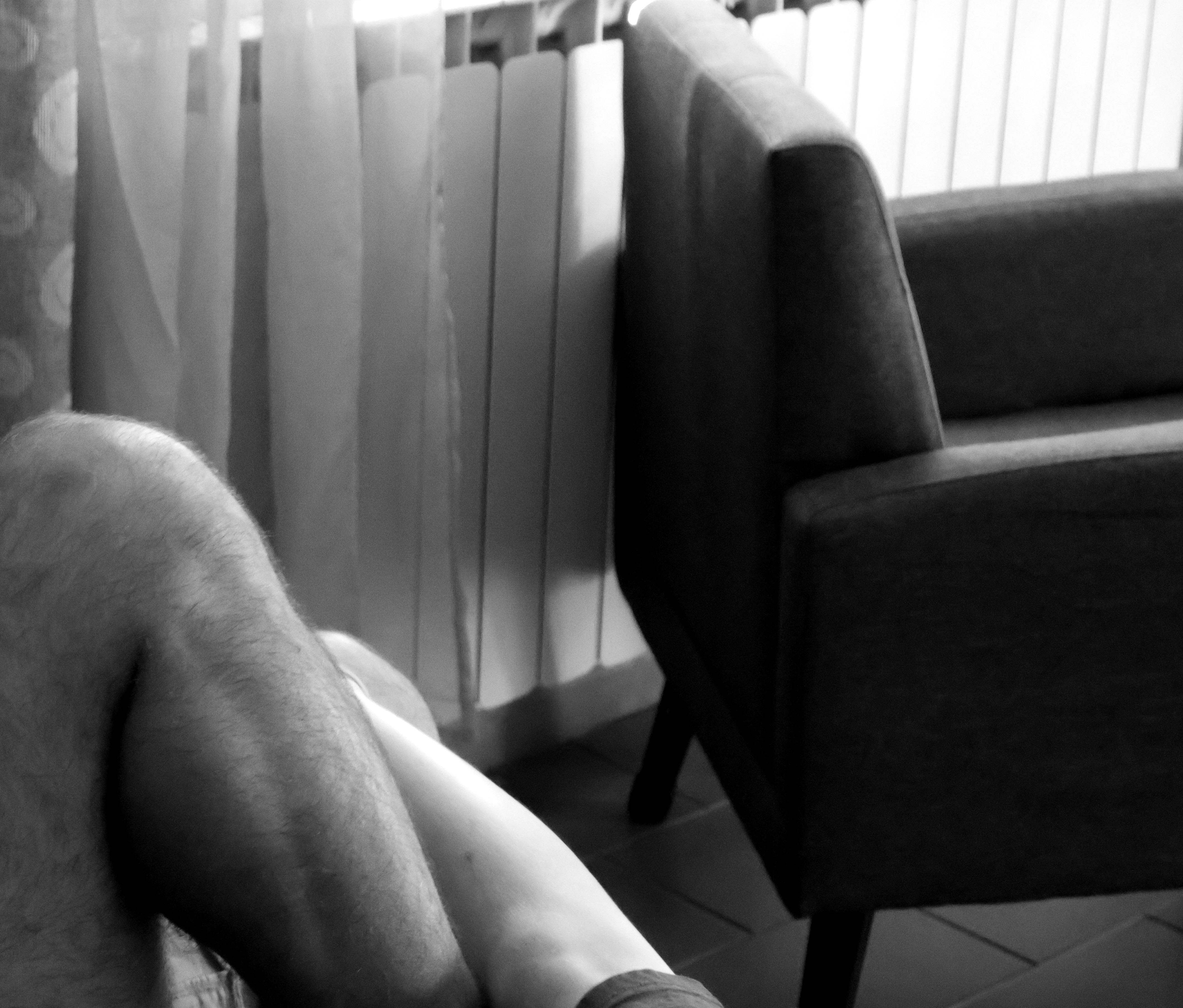Gambe vicine in primo piano accanto a una poltrona e a una finestra con tende, in bianco e nero
