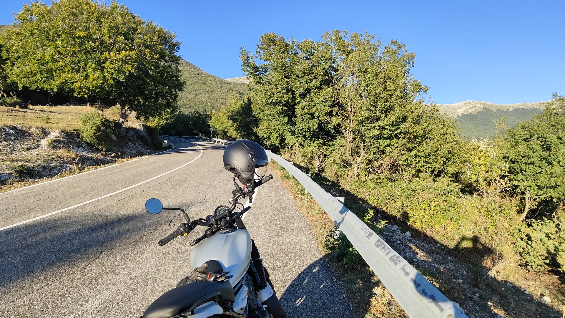 Una moto ferma lungo una strada di montagna, con il casco sul manubrio, alberi e cielo azzurro