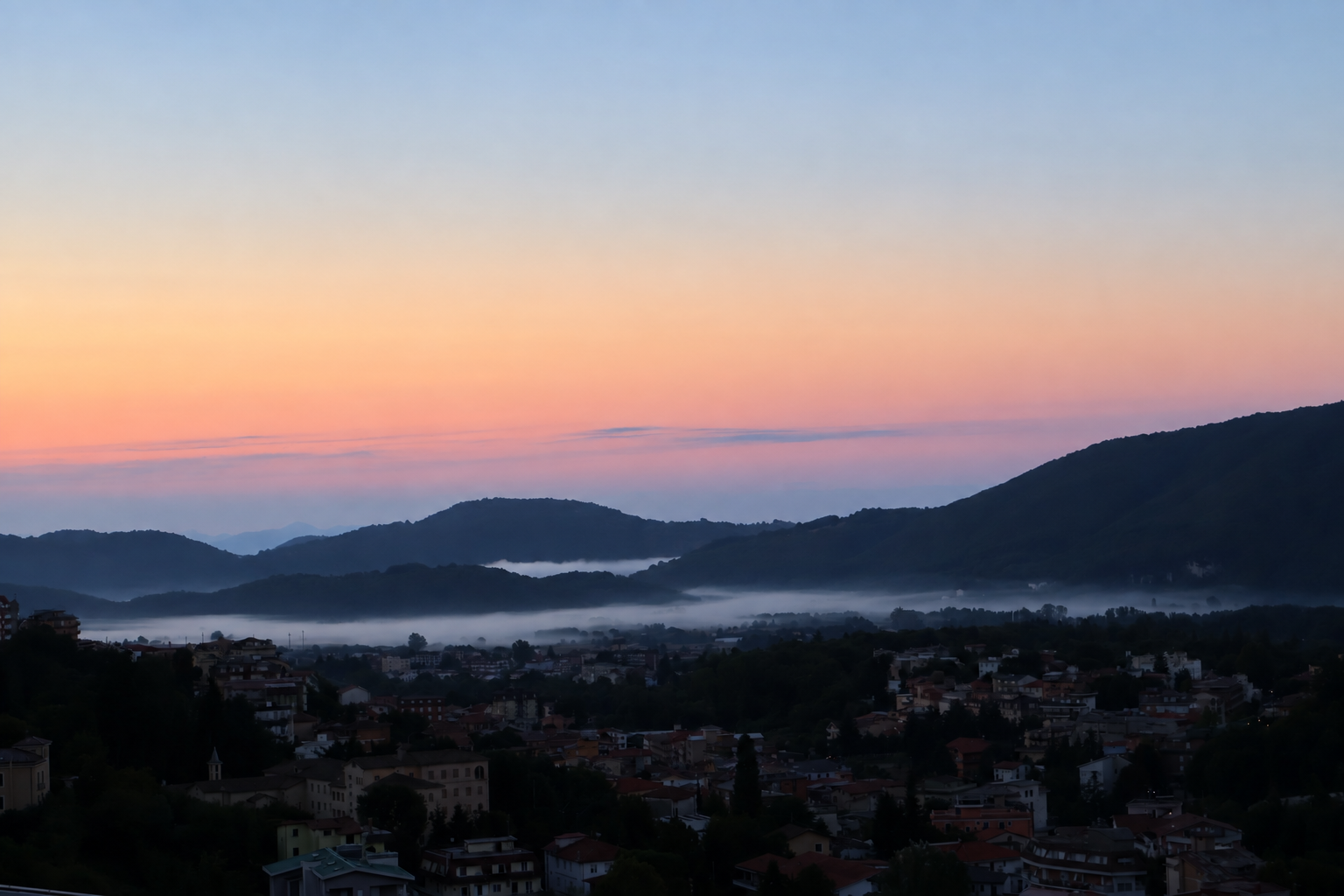 Il paese ancora nell'ombra, la nebbia tra le colline e il cielo rosa del mattino